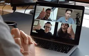 A person is viewing a laptop screen displaying a video call with four people. Three of them are giving a thumbs-up.